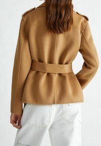 Kurtka z mieszanki wełny wielbłądziej o dopasowanej talii, szerokim pasku, pagonami na ramionach i gładkiej fakturze. Poniżej widoczne białe dżinsy.