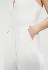 Bílý elegantní overal s hlubokým výstřihem do V, širokým pasem, sklady na přední straně a bočními kapsami, vyrobený z hladkého, lehkého materiálu.