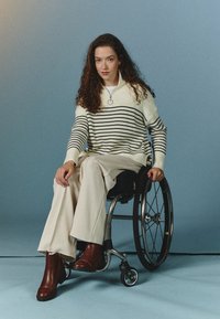 Strikkegenser med sorte striper på en kremfarget bakgrunn, sammen med høylivede beige bukser og brune ankelstøvletter. Rullestol synlig.