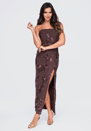 Femme posant dans une robe bustier marron foncé à fleurs avec une fente haute sur la cuisse et des talons ouverts dorés sur un fond clair uni.