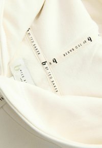 Crèmekleurige stof met een zachte textuur, voorzien van een etiket met de tekst "b by Ted Baker" en een kleinere maatlabel. Minimalistisch ontwerp.