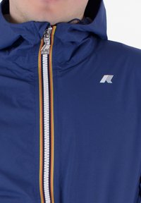 Giacca blu con zip e cappuccio. Presenta una zip a contrasto con arcobaleno e un piccolo logo sul petto. Realizzata in tessuto liscio e leggero.