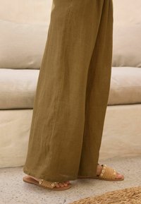 Olivegrüne Leinen-Hosen mit weitem Bein, kombiniert mit tan-farbenen, geflochtenen Riemchensandalen mit Muscheln. Der strukturierte Stoff kontrastiert mit glatten Oberflächen.