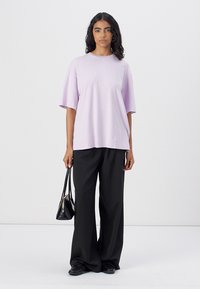 Tricou din bumbac de culoare violet deschis, cu o croială lejeră, mâneci scurte și guler de tip crew, asortat cu pantaloni negri cu croială largă și o geantă mică neagră.