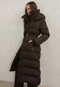 Piumino marrone scuro con collo alto, vita con cintura e tasche con cerniera; presenta un design trapuntato a linee verticali e una silhouette lunga.