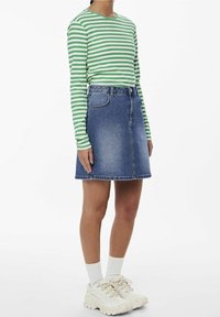 Henkilö, jolla on päällään vihreä-valko-raidoitettu pitkähiha topi, siniset farkkuskortit, valkoiset sukat ja paksut valkoiset tennarit, seisoo valkoista taustaa vasten.