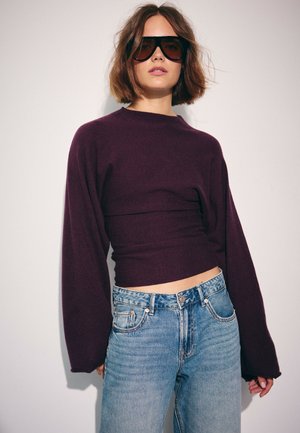 Femme aux cheveux bruns courts portant de grandes lunettes de soleil noires, un haut ajusté à manches longues violet foncé et un jean taille haute bleu clair.