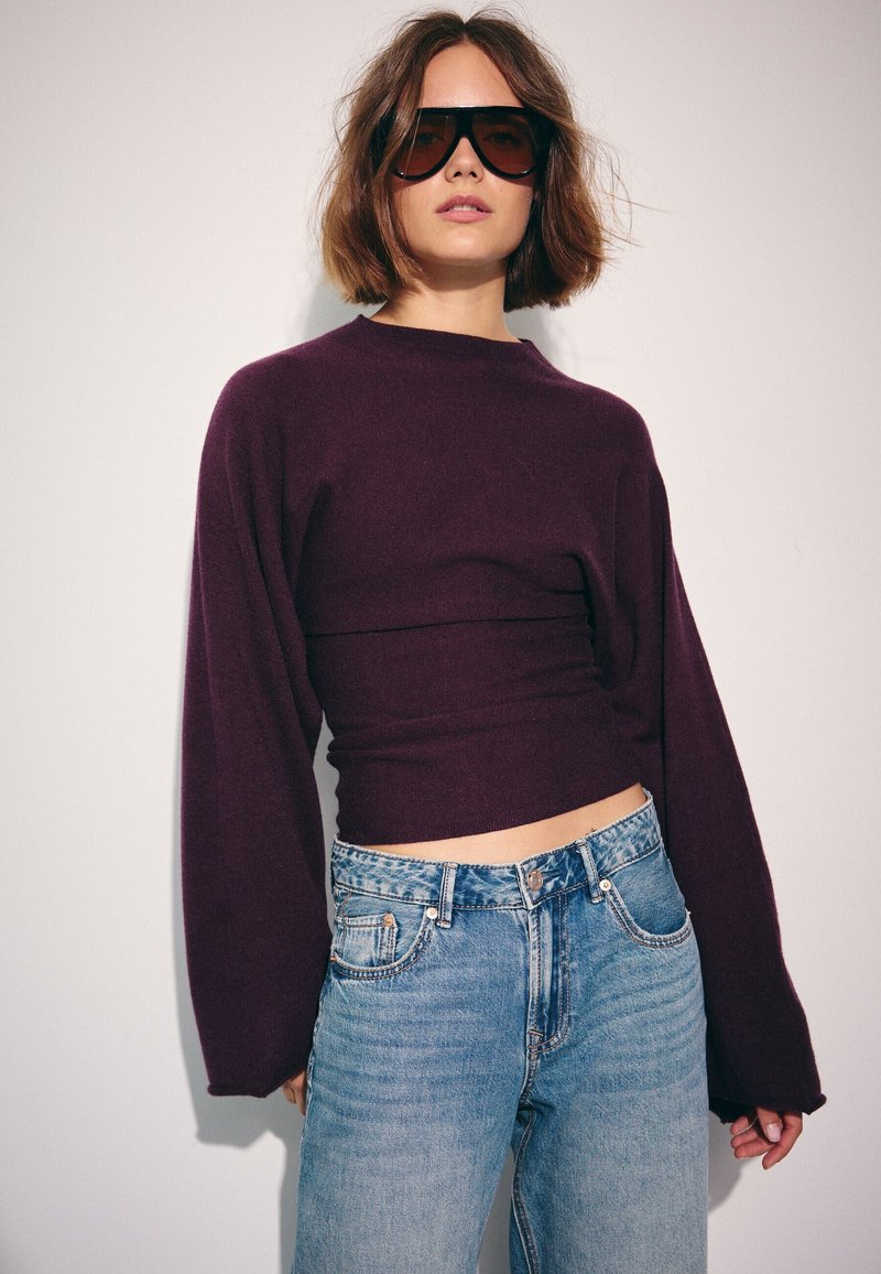 Femme aux cheveux bruns courts portant de grandes lunettes de soleil noires, un haut ajusté à manches longues violet foncé et un jean taille haute bleu clair.
