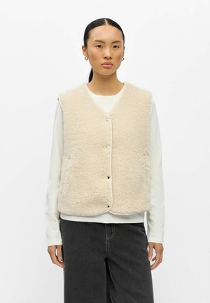 Jeune femme aux cheveux foncés attachés porte un gilet beige duveteux sur une chemise blanche à manches longues et un pantalon gris foncé, debout sur un fond blanc.