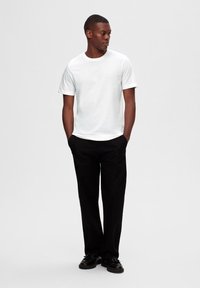 Camiseta de algodón blanca con mangas cortas y cuello redondo, combinada con pantalones negros y zapatos negros, de pie contra un fondo neutro.