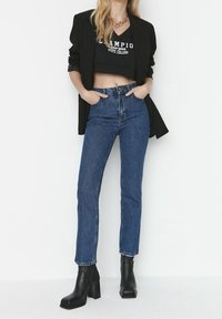 Jeans blu a vita alta con una texture liscia e una leggera sfumatura, abbinati a un top nero corto e a un blazer nero, insieme a stivaletti con tacco grosso.