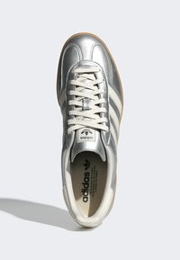 Silver Adidas-sneaker med vita ränder, vita snörningar, gummisulor och svart Adidas-logotyp på tungan och innersulan, visad uppifrån.