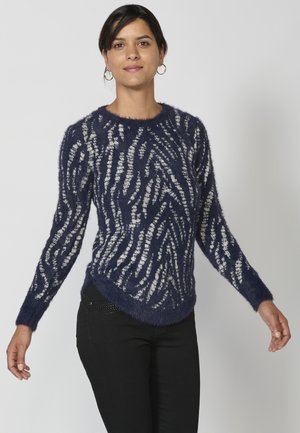 Marineblauer, flauschiger Pullover mit einem strukturierten weißen Wellenmuster, langen Ärmeln und abgerundetem Saum. Kombiniert mit schwarzen Hosen.