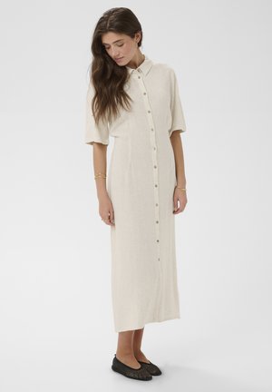 Kobieta ubrana w beżową, zapinaną na guziki sukienkę midi z krótkimi rękawami, stojąca i patrząca w dół, w połączeniu z czarnymi plecionymi balerinami i złotymi bransoletkami.