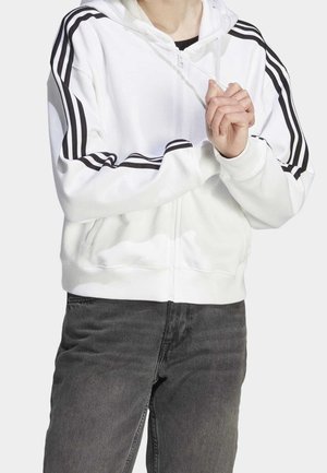 Person iført hvid hættetrøje med lynlås og sorte striber på ærmerne, holder i snørebåndene på hættetrøjen, kombineret med mørkegrå denimjeans.