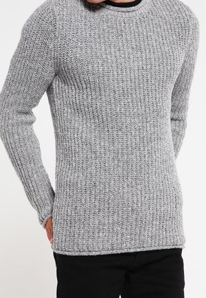 Nahaufnahme einer Person, die einen grau gestrickten Pullover mit gerippter Struktur und schwarze Hosen trägt, Hände sind teilweise an den Seiten sichtbar.