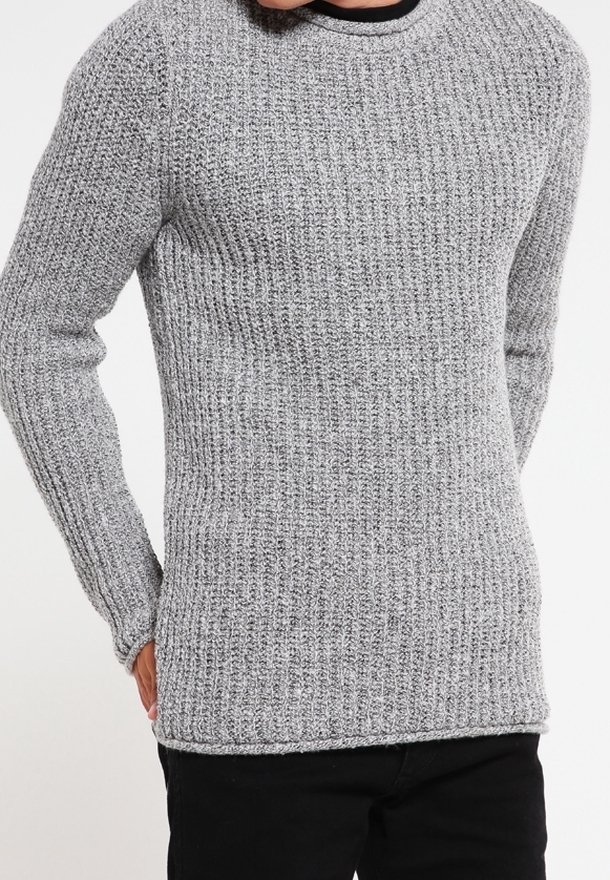 Gros plan d'une personne portant un pull gris tricoté avec une texture côtelée et un pantalon noir, les mains partiellement visibles sur les côtés.