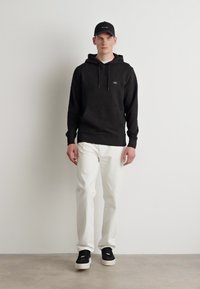 Hanorac negru cu glugă și șnururi, pantaloni albi și adidași negri. Modelul stă în fața unui zid simplu, evidențiind o ținută casual.