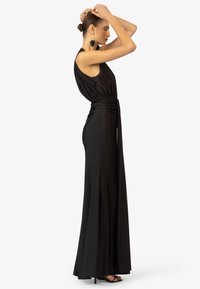 Schwarzes ärmelloses Maxikleid mit drapierter Bodice, tailliertem Schnitt, fließendem Rock und seitlichem Schlitz. Begleitet von großen Ohrringakzenten.
