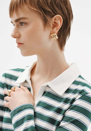 Camisa polo a rayas verdes y blancas con un cuello blanco, y un modelo que lleva pendientes de aro dorados y múltiples anillos, mostrando un perfil cercano.