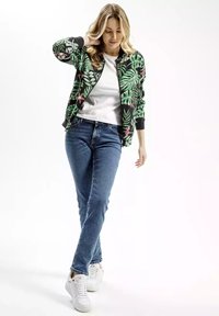 Kwiatowa kurtka bombowa z różowymi i zielonymi wzorami liści na czarnym tle, zestawiona z białą koszulką i niebieskimi dżinsami, białe sneakersy.