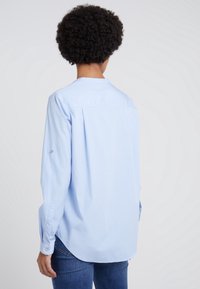 Jasnoniebieska koszula zapinana na guziki z długimi rękawami, zaokrąglonym dołem i detalem w postaci zakładki z tyłu. Miękki, oddychający materiał.