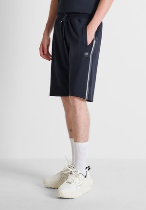 Uomo che indossa pantaloncini neri casual con banda laterale, calzini atletici bianchi e sneakers bianche chunky, in piedi con una mano in tasca.