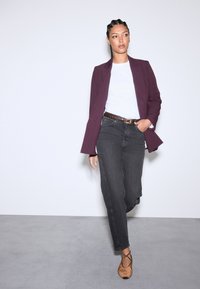 Dunkelgraue Jeans, weißes T-Shirt und ein bordeauxroter Blazer. Der Blazer hat ein strukturiertes Design; die Schuhe sind hellbraun mit gekreuzten Riemen.