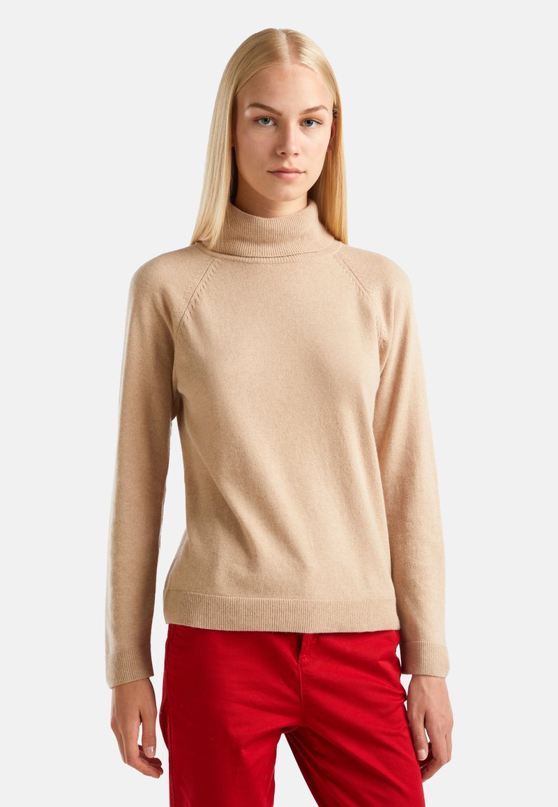 Jeune femme aux cheveux blonds raides portant un pull à col roulé beige et un pantalon rouge, se tenant devant un fond blanc uni.