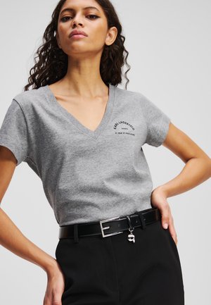 T-shirt en coton gris à col en V avec des manches courtes, arborant un logo imprimé noir. Associé à une ceinture noire avec une boucle en argent et un pendentif.