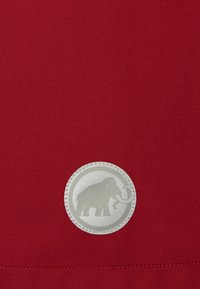 Rødt stof med en glat tekstur har et rundt, sølvfarvet logo, der forestiller en mammut, fremhævet med en syet kant.