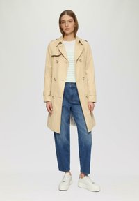 Beige trenchcoat met dubbelzijdige knopen, gecombineerd met denim jeans en witte sneakers. Lichte gestreepte top zichtbaar eronder.