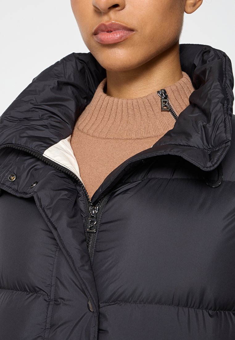 Schwarze Pufferjacke mit hohem Kragen, Reißverschluss und dekorativen Beschlägen. Die Details umfassen einen weichen beigen Rollkragenpullover darunter.