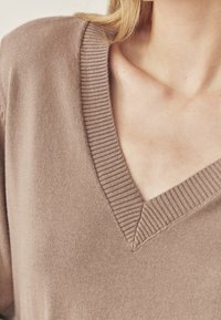 V-veida kakla džemperis no mīksta, gaiši brūna trikotāžas auduma. Raksturo ribota apkakle un tekstūra ar vaļīgu piegriezumu. Tuva skata attēls izceļ auduma detaļas.