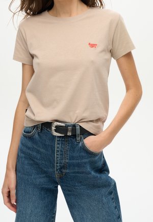 Beige cotton t-shirt with short sleeves, featuring a red logo on the left chest pocket area, paired with high-waisted blue denim jeans and a black belt.
