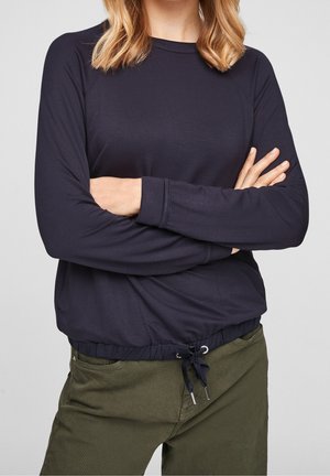 Personne aux cheveux blonds portant un haut bleu marine à manches longues avec cordon à la taille et un pantalon vert olive, les bras croisés sur la poitrine.