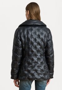 Black puffer jacket with quilted design, featuring a large collar and textured surface. Model in denim jeans shows the back view.