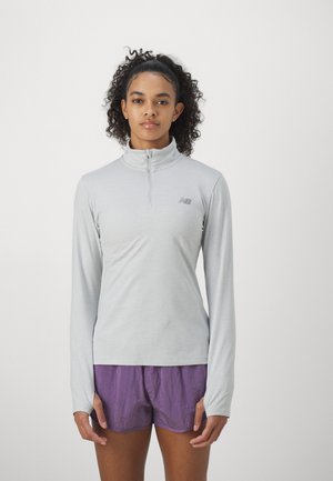 Light gray long-sleeve athletic top with a zippered collar, featuring a logo on the left chest. Paired with purple shorts. Smooth texture.