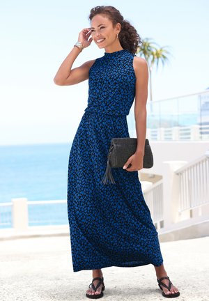 Maxikleid mit einem blau-schwarzen Punktenmuster, ärmellosem Design und taillierter Taille. Kombiniert mit einer strukturierten schwarzen Clutch und Sandalen.