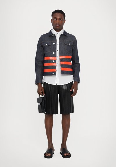 Veste en denim avec des bandes réfléchissantes orange, chemise blanche à boutons, short plissé noir et sandales noires. Le modèle tient un petit sac.