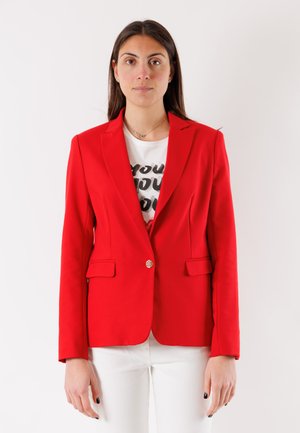 Donna con lunghi capelli scuri che indossa un blazer rosso acceso sopra una t-shirt bianca con grafica e pantaloni bianchi su uno sfondo chiaro e uniforme.
