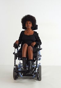 Cadeira de rodas elétrica preta com um encosto de apoio e recurso de reclinação. Pessoa vestida com um vestido preto justo e botas altas pretas sentada.