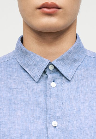 Κοντινό πλάνο του κάτω μέρους του προσώπου και του λαιμού ενός ατόμου που φοράει ένα ανοιχτό μπλε λινό πουκάμισο με κουμπιά και κλασικό γιακά.