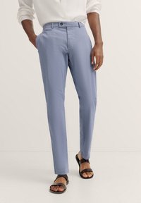 Hellblaue Hose aus strukturiertem Stoff, mit flachem Bund, seitlichen Taschen und schwarzem Knopfverschluss, kombiniert mit schwarzen Sandalen.