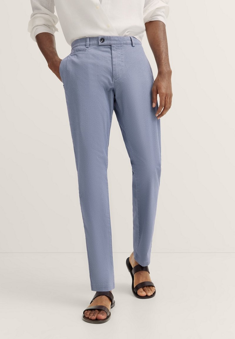 Hellblaue Hose aus strukturiertem Stoff, mit flachem Bund, seitlichen Taschen und schwarzem Knopfverschluss, kombiniert mit schwarzen Sandalen.