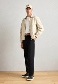 Jachetă bej ușoară, tricou alb, pantaloni negri, adidași gri cu accente portocalii și o căciulă bej deschis. Stând pe podea din lemn.