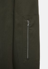 Groene stof met een gladde textuur, voorzien van een ritszak aan de zijkant. De rits is metallic met een kleine trekhaak.