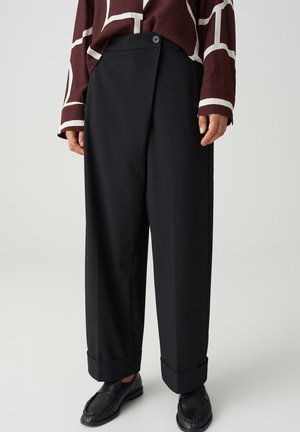 Persona che indossa pantaloni neri a gamba larga con orli risvoltati, abbinati a mocassini neri e una maglia marrone a maniche lunghe con linee geometriche bianche.