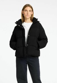 Sort pufferjakke med hætte, trykknapper og sidelommer. Designet er quilted med en glat tekstur, båret over en hvid skjorte.