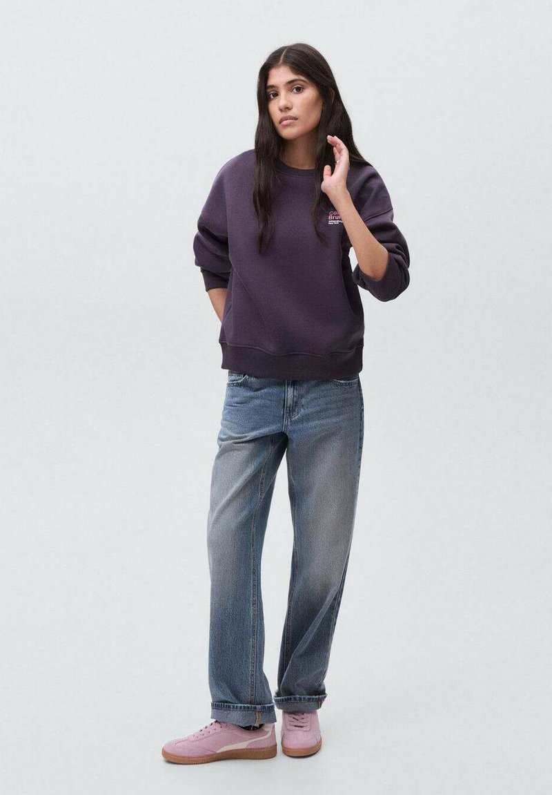 Kvinna med långt mörkt hår som bär en lila sweatshirt, lösa blå jeans och rosa sneakers, stående mot en enkel ljus bakgrund.